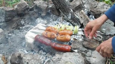 Brochettes sosis ile yuvarlak dönüm eller görünümü kapatın. Dört sosis ve soğan bahçesinde sıcak Yanan köz üzerinde ızgara ile tükürüyor. Ateş çukuru taşlarla döşeli içinde sigara köz.