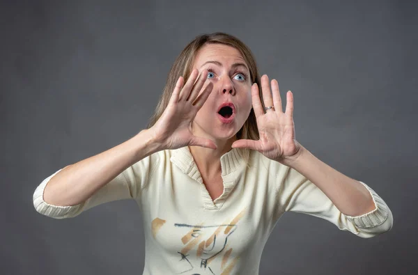 Emozioni femminili. Una giovane donna urla ad alta voce a qualcuno. Ritratto di ragazza con le mani in bocca. Linguaggio del corpo e sentimenti reali. Ritratto di donna emotiva su sfondo grigio ripresa in studio . — Foto Stock