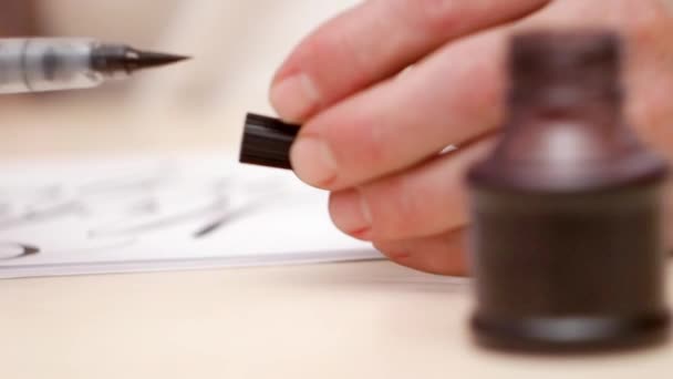 Mans hands close brush pen then put pen down on beige desk. Close shot. Front view. Calligraphic lettering. Open inkpot on beige table. Calligraphic tools. Selective soft focus. Blurred background. — Stock Video