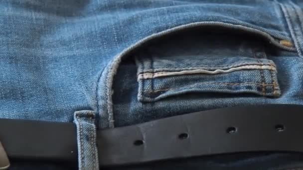 Blue denim jeans with leather belt laying on table, close shot. Macro dolly shot. Selective soft focus. Camera moving along belt and front trouser pockets — Stock Video