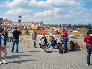 Prag, Çek Cumhuriyeti - Nisan 2019: Charles Köprüsü'nde çalan sokak müzik grubu. Charles Köprüsü'nde yürüyen ve sokak bandını dinleyen turistler. Saksafonçalan adam. Prag'da bulutlu bir gün.