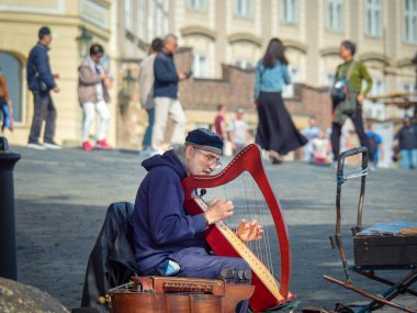 Prag, Çek Cumhuriyeti - Nisan 2019: Charles köprüsünde oturan ve arp çalan yaşlı adam. Arpıyla sokak müzik oyuncusu. Prag'da köprüde yürüyen turistler, bulanık arka plan.