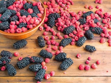 Taze sulu meyveler kahverengi masaya yığılmış, yakından görünümü. Olgun kırmızı ahududu ve kara dut dolu kahverengi kase. Ahşap masa dokusu. Seçici yumuşak odak. Bulanık arka plan.