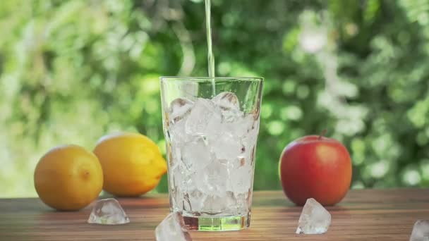 Rociador de agua vertiendo en un vaso lleno de cubitos de hielo, tiro cerrado. Limones amarillos, manzana roja y cubitos de hielo en el escritorio marrón. Hojas verdes sobre fondo. Enfoque suave selectivo. Fondo borroso . — Vídeos de Stock