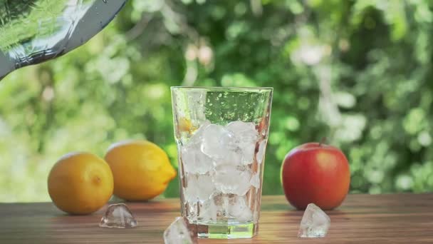 Mesa de agua vertiendo en un vaso lleno de cubitos de hielo, tiro cerrado. Manzana roja, limones amarillos y cubitos de hielo en el escritorio marrón. Hojas verdes balanceándose en el viento. Enfoque suave selectivo. Fondo borroso . — Vídeo de stock