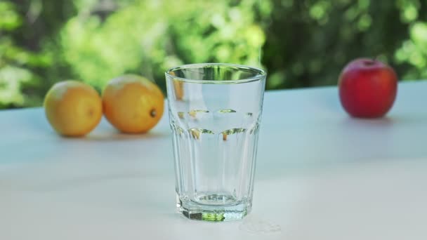 Derretimiento de cubos de hielo lanzando en un vaso vacío, tiro cerrado. Limones amarillos, manzana roja y vidrio sobre superficie blanca. Greenth en segundo plano. Enfoque suave selectivo. Fondo borroso . — Vídeos de Stock