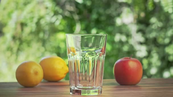 Cubos de hielo cayendo en un vaso vacío, tiro cerrado. Limones amarillos, manzana roja y vidrio sobre escritorio marrón. Hojas verdes balanceándose en el viento sobre el fondo. Enfoque suave selectivo. Fondo borroso . — Vídeos de Stock