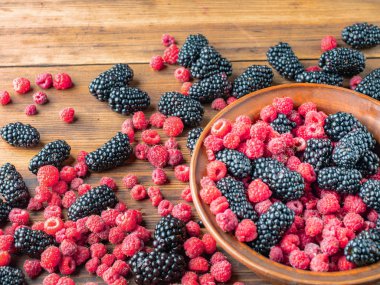 Kırmızı ahududu ve siyah dut kahverengi masaya yığılmış, yakın görünümü. Olgun meyvelerle dolu kahverengi kase. Ahşap masa dokusu. Seçici yumuşak odak. Bulanık arka plan.