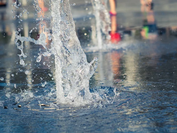 Close up view on water streams flowing out of stainless grate of dry fountain. Kids bare feet cooling near fountain. Town square in summer. Selective soft focus. Blurred background.