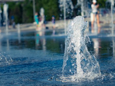 Kuru çeşmenin granit kaplamasına sıçrayan dikey su akışları, görünümü kapatın. Çeşmenin içinde soğutan çocuklar. Yaz şehir manzarası. Seçici yumuşak odak. Bulanık arka plan.