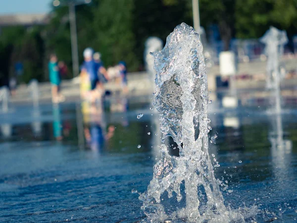 Dry fountain, close up view. Vertical water stream flowing out of stainless grate of fountain. Kids walking bare feet inside fountain. Town square in summer. Selective soft focus. Blurred background. — Stock Photo, Image
