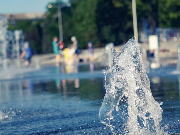 Kuru çeşme dikey su akışları, yakın görünümü. Fıskiyenin yanında soğuyor çocukları olan ebeveynler. Şehir meydanı, yaz manzarası. Seçici yumuşak odak. Bulanık arka plan.