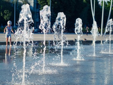 Kuru çeşmedikey su akar, yakın görünümü. Fıskiyenin yanında yürüyen bir çocuk çıplak ayak. Granit kaplamaüzerine sıvı sıçraması. Yaz şehir manzarası. Seçici yumuşak odak. Bulanık arka plan.