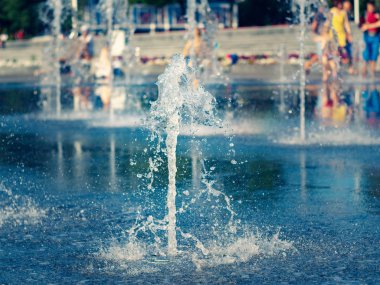 Granit kare kaplamadan akan su akışlarının görünümünü kapatın. Kuru çeşmenin yanında soğuyan ebeveynleri olan çocuklar. Yere su sıçratıyor. Seçici yumuşak odak. Bulanık arka plan.