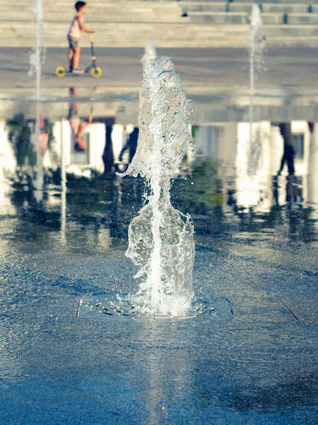 Fuente seca. Vista de cerca de la corriente vertical de agua que sale de la rejilla de acero inoxidable. Chico conduciendo patinete scooter. Paisaje urbano de verano. Enfoque suave selectivo. Fondo borroso . —  Fotos de Stock