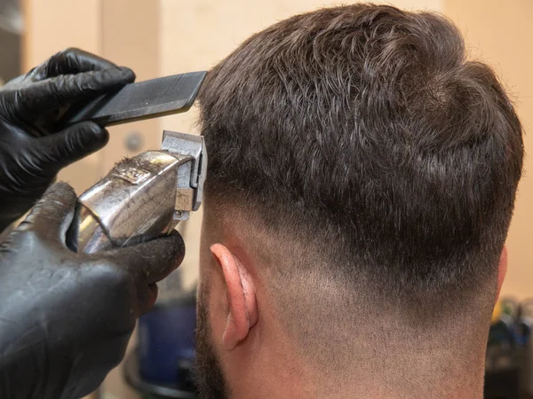 Hairstylist working with clipper and comb, close up view. Mans head and masters hands in black rubber gloves. Stylist at work in studio. Selective soft focus. Blurred background. — Stock Photo, Image