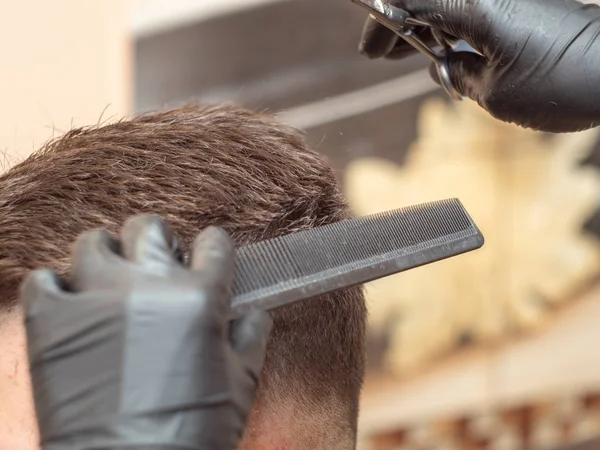 Close up on males hair in barbershop. Hairstylists hands in black rubber gloves. Master cutting hair with comb and scissors. Master and client in barbershop. Selective soft focus. Blurred background.