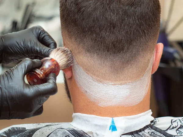 Hairstylist spreading shaving foam with shaving brush on clients neck, close up view. Hands in black rubber gloves and males nape close-cropped. Selective soft focus. Blurred background. — Stock Photo, Image