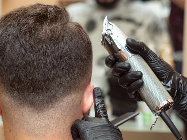 Coupe de cheveux avec tondeuse, nuque masculine, vue rapprochée. Mains stylistes en gants de caoutchouc noir avec tondeuse. Faire une coiffure moderne dans le salon de coiffure. Vue rapprochée. Fond flou . — Photo