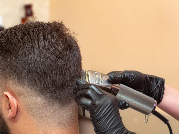 Stilist erkekler ense üzerinde saç kesme makas ile, yakın görünümü. Stilistler elleri siyah lastik eldivenler. Modern saç modeli yapıyorum. Berber dükkanının bej duvarı. Seçici yumuşak odak. Bulanık arka plan.
