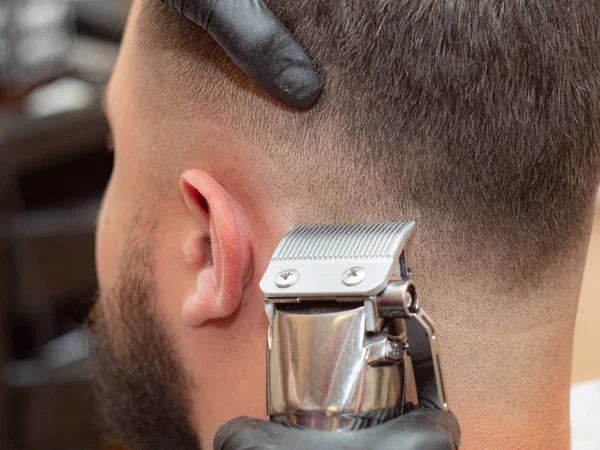 Hårklippning process, fotograferad i barbershop, närbild vy. Stylist klippa hår med Clipper. Masters händer i svarta gummihandskar. Selektivt mjukt fokus. Suddig bakgrund. — Stockfoto