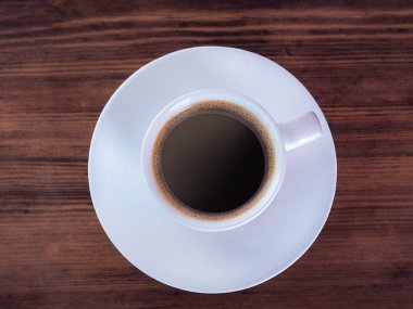 Beyaz fincan aromatik kahve dolu, yakın, üst görünümü. Beyaz kahve fincanı ve kahverengi masa üzerinde tabak. Ahşap masa dokusu. Seçici yumuşak odak. Bulanık arka plan.