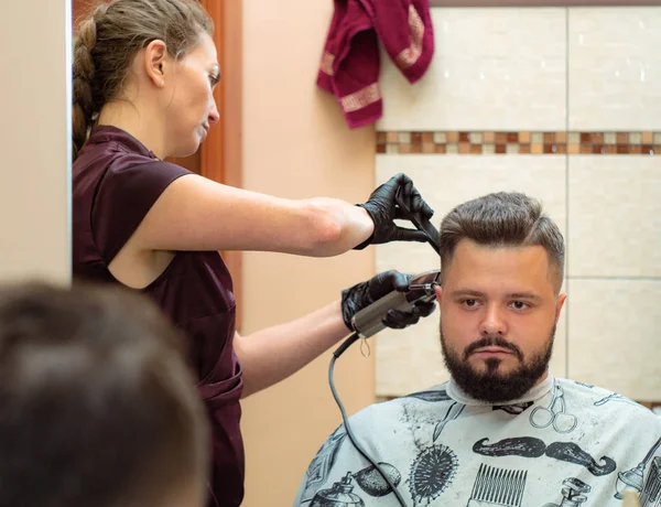 Jovem mulher trabalhando com cortador e pente, vista de perto. Mãos em luvas de borracha preta. Estilista e cliente refletindo no espelho. Fotografado na barbearia. Foco suave seletivo. Fundo desfocado . — Fotografia de Stock