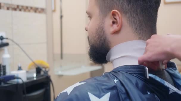 Homme avec barbe assis sur une chaise dans un tablier. Coiffeur mis sur bande autour du cou du client au salon de coiffure. Fond flou, fond de mise au point douce. Vue rapprochée de l'arrière — Video