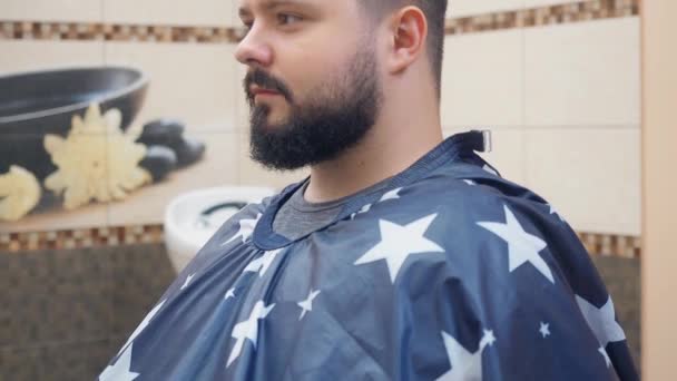 Hairdresser comes and put on paper tape around the neck of the client at the Barbershop. Man with beard sitting on chair in an apron. Middle plan from side. Blurred background, soft focus. — Stock Video