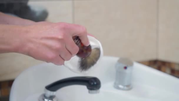 Stylist preparing shaving foam with brush above white washstand, close shot. Recorded in hairdressing saloon. Interior of barbershop. Selective soft focus. Blurred background. — Stock Video