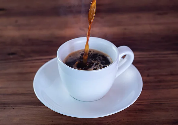 Koffie gieten in witte beker, close-up weergeven. Koffie kopje vol aromatische espresso. Witte kop en schotel op houten tafel. Selectieve soft focus. Onscherpe achtergrond. — Stockfoto