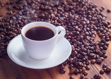 Kahve çekirdekleri yığını ve kahverengi masanın üzerinde siyah espresso dolu beyaz fincan, yakın görüş. Kahverengi masa dokusu. Seçici yumuşak odaklanma. Bulanık arkaplan.