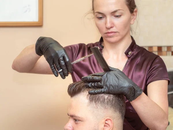 年轻的女性用剪子和黑色的梳子剪客户的头发，近距离观察。 设计师手戴黑色橡胶手套. 理发店的内部 有选择的软焦点。 模糊的背景. — 图库照片