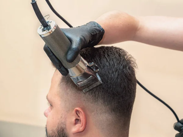 Cabeleireiro trabalhando com clipper, vista de perto. Processo de corte de cabelo. Mãos de mestre em luvas de borracha preta. Fotografado na barbearia. Foco suave seletivo. Fundo desfocado . — Fotografia de Stock