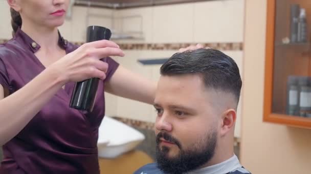 Estilista en vestido rojo oscuro fijando clientes peinado con spray para el cabello y peine, tiro cerrado. Grabado en barbería. Interior del salón de peluquería. Enfoque suave selectivo. Fondo borroso . — Vídeos de Stock