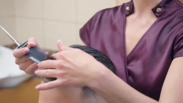 Stylist cutting mans hair with comb and scissor, close shot. Female making modern hairdo. Recorded in barbershop. Selective soft focus. Blurred background. — Stock Video
