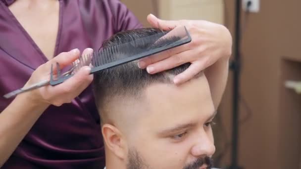 Male in hairdressing saloon, close shot. Female in dark red dress cutting hair with scissor and comb. Master at work in barbershop. Selective soft focus. Blurred background. — Stock Video