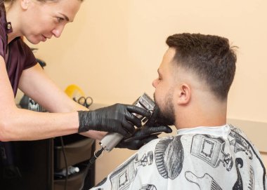 Genç bir bayan, kırpıcı ve tarak ile müşterileri kesiyor, manzarayı kapatın. Siyah lastik eldivenli eller. Berber dükkanında bir erkek. Kuaför salonunun içi. Seçici yumuşak odaklanma. Bulanık arkaplan.