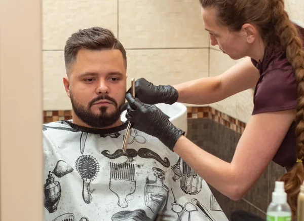 Jovem fêmea barbear machos castor com lâmina aberta, vista de perto. Estilistas mãos em luvas de borracha preta. Masculino e estilista refletindo no espelho. Foco suave seletivo. Fundo desfocado . — Fotografia de Stock