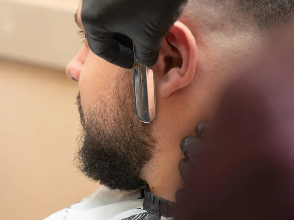 Estilista barbear machos castor com lâmina aberta, vista de perto. Mãos de mestre em luvas de borracha preta. Fotografado na barbearia. Foco suave seletivo. Fundo desfocado . — Fotografia de Stock