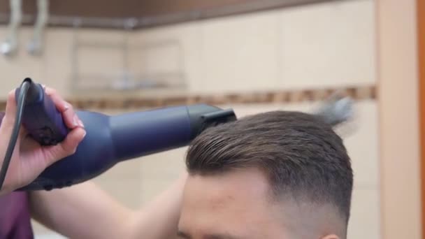 Estilista haciendo peinado moderno con secador de pelo y peine redondo, tiro cerrado. Hombre en peluquería. Interior de la barbería. Enfoque suave selectivo. Fondo borroso . — Vídeos de Stock