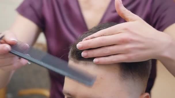 Close shot van stylisten handen knippen haar met kam en schaar. Een jongeman in een kapsalon. Op bezoek bij de kapper. Selectieve zachte focus. Wazige achtergrond. — Stockvideo