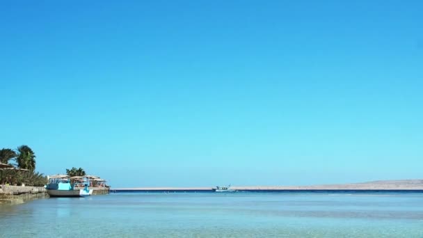 Playa del Mar Rojo con muelle, tiro ancho extremo. Un barco atado a un muelle otro navegando por el mar. Cielo azul despejado. Fotografiado en Hurghada en febrero. Enfoque suave selectivo. Fondo borroso . — Vídeos de Stock