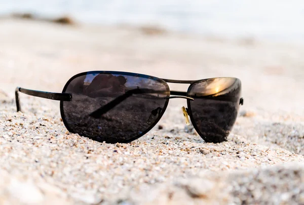 Солнцезащитные очки на песке с отражением заката на море и фотограф — стоковое фото