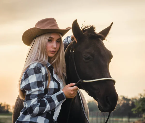 Mladá dívka oblečená v kovbojském klobouku a kostkované košili s koněm při západu slunce — Stock fotografie