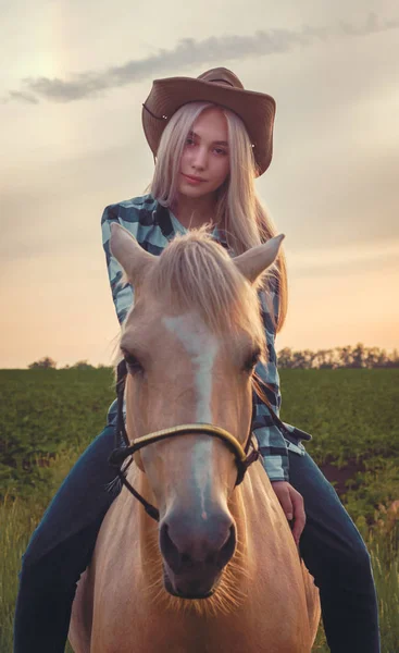 Niña vestida con un sombrero de vaquero y vaqueros azules se sienta en un caballo contra el telón de fondo de la puesta de sol — Foto de Stock