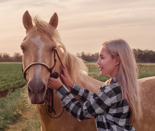 Chica rubia joven con un caballo beige en el rancho en el fondo del campo y el cielo de la noche — Foto de Stock