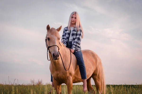 年轻的金发女孩穿着蓝色牛仔裤和格子衬衫坐在马在牧场特写 — 图库照片