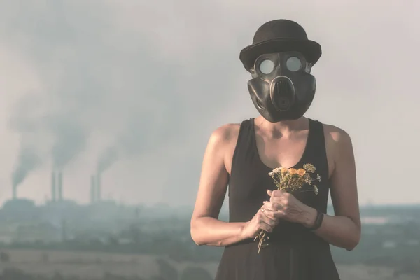 Jong meisje in een zwarte jurk en een gasmasker met bloemen op de achtergrond van roken fabriek schoorstenen — Stockfoto