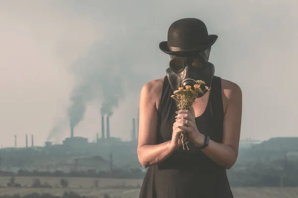 Young girl in a black dress and a gas mask with flowers on the background of smoking factory chimneys — Stock Photo, Image